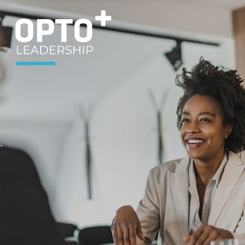 A professional woman with curly hair, wearing a light-colored blazer, is engaged in a conversation with another person in a business setting. She is smiling and appears confident. The "OPTO+ LEADERSHIP" logo is visible in the top left corner.